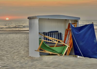 houten strandtent met gekleurde vouwstoelen en blauwe windscherm bij neergaande zon boven zee
