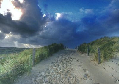 strandopgang met voetsporen in het zand en prikkeldraad en grashalmen aan beide kanten met zicht op horizon en dreigende wolken