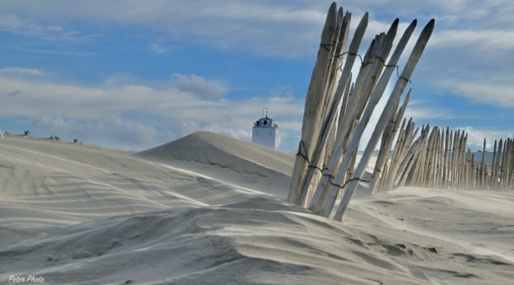 stuivend zand achter een hek van palen op de voorgrond met de top van de vuurtoren katwijk zichtbaar achter het duin